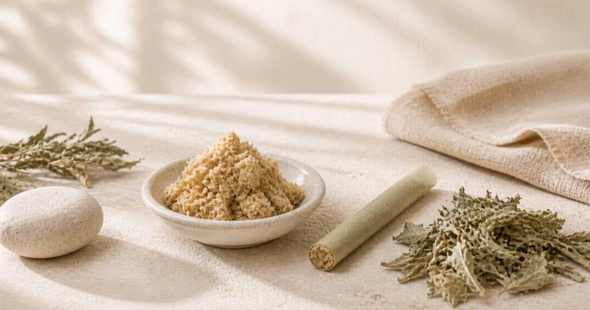 A bowl of moxa floss, a moxa stick, and dried mugwort sprigs arranged on cream linen
