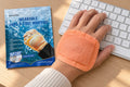 A peach-orange warmer on the back of a hand using a keyboard, beside a blue pouch.