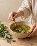 An illustrative historical scene of hands stirring herbs into oil in an earthenware bowl.